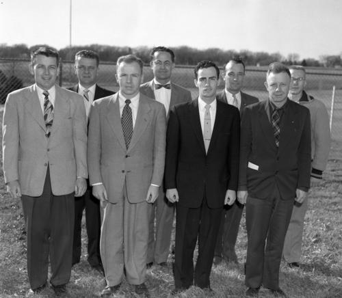 Group of eight male Bootstrapper graduates standing for a photo outside in front of a field at the University of Omaha in 1957., UNO Libraries' Archives &amp; Special Collections
