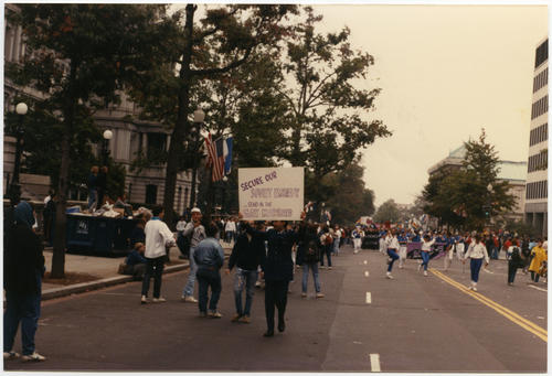 Unidentified probable Caucasian person in military dress uniform (dark blue) walking in parade route and holding handmade sign high above their head, "Secure our Soviet Embassy...Send in the Gay Marines" in Washington D.C. October 11, 1987 at the Second National March on Washington for Lesbian and Gay Rights., UNO Libraries' Archives &amp; Special Collections